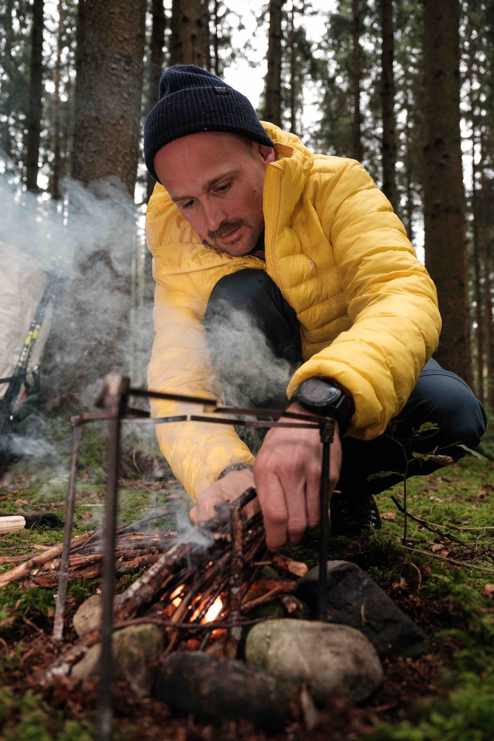 Manuel Seiler der Outdoorguide entfacht ein Feuer mit kleinen Ästen mitten im Moosgrünen Wald und trägt eine blaue Mütze